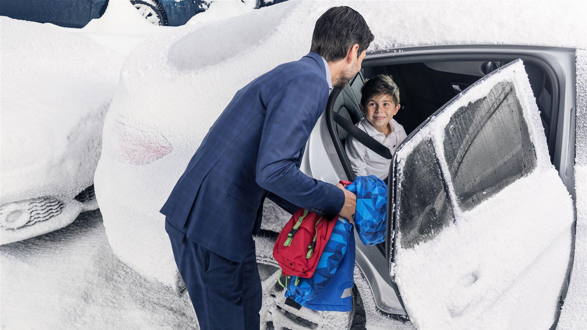 Kind sitzt im eingeschneiten Auto und Vater reicht warme Sachen durch Tür