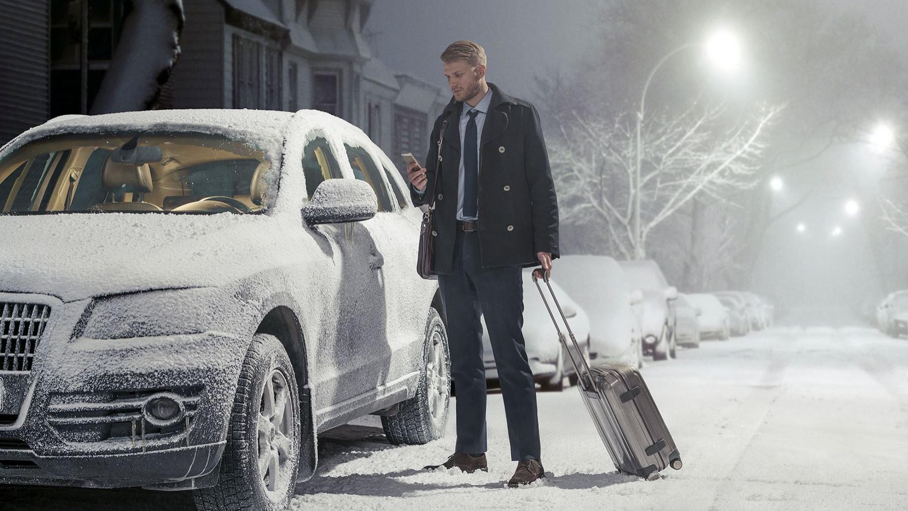 Businessmann steht mit Smartphone am eingeschneiten Auto, nur die Scheiben sind abgetaut