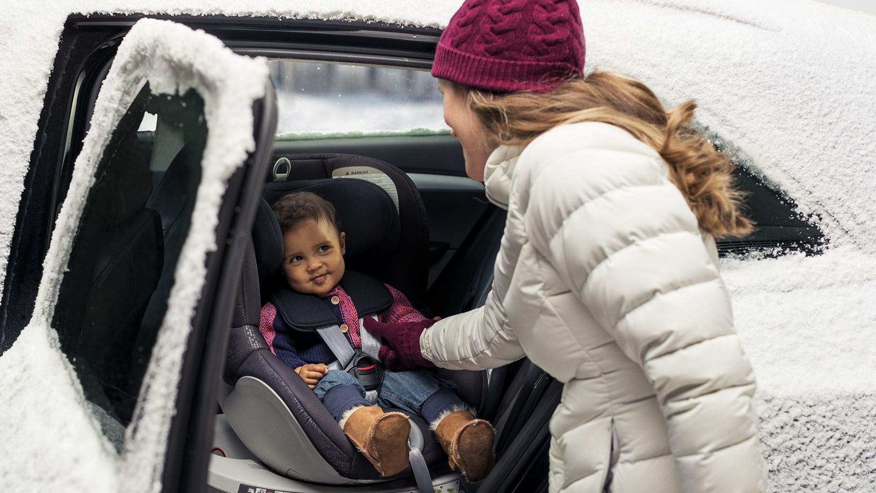 Mutter setzt Kleinkind in Kindersitz auf Rücksitzpank im eingeschneiten Auto
